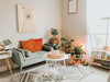 A cozy, well-lit living room with a gray sofa, orange cushion, white coffee table, plants, a rug, and a hanging inspirational quote poster.